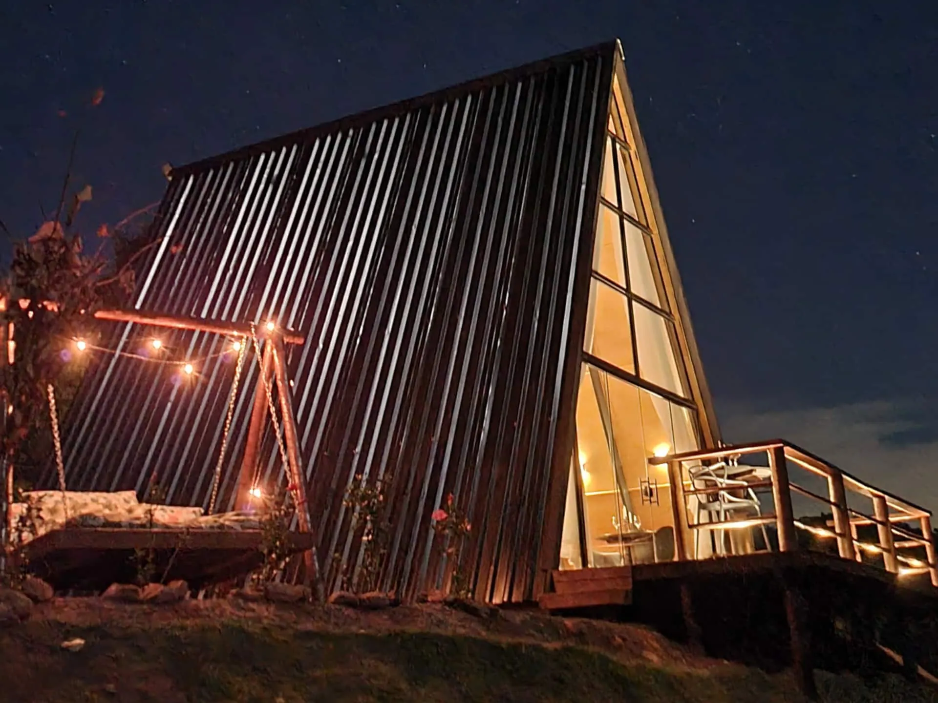 O chalé A-frame iluminado à noite, com o deck e o varal de luzes